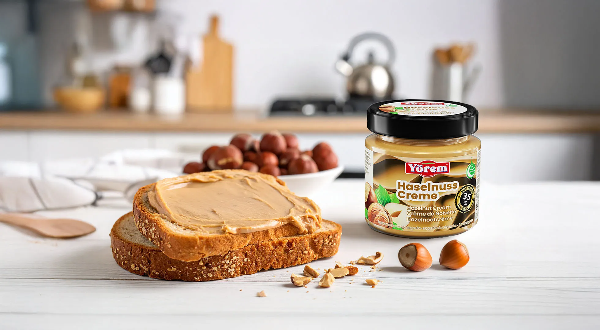 Ein Glas „Yörem Haselnuss Creme“ steht in einer Küche auf einem Tisch. Vor dem Glas liegt eine Scheibe Vollkornbrot, dick bestrichen mit der cremigen Haselnusscreme. Daneben sind ganze und aufgebrochene Haselnüsse verstreut. Auf dem Etikett des Glases ist der Produktname „Haselnuss Creme“ mit dem Hinweis „35 % Haselnüsse“ sichtbar.