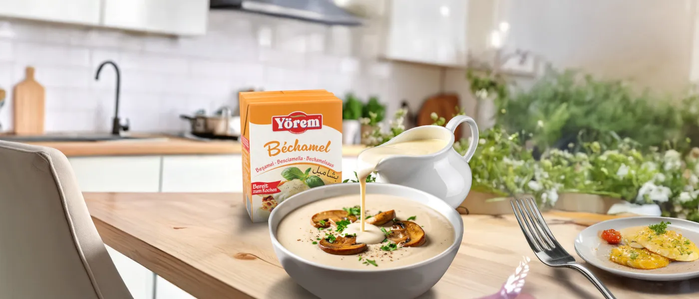 Eine orangene Packung Yörem Béchamelsauce steht auf einem Küchentisch in einer modernen, hellen Küche. Daneben wird cremige Béchamelsauce aus einem weißen Krug in eine Pilzcremesuppe gegossen. Im Vordergrund steht ein Teller mit Ravioli, im Hintergrund ist ein Fenster mit Blick ins Grüne zu sehen. Die Szene wirkt frisch, einladend und appetitlich.