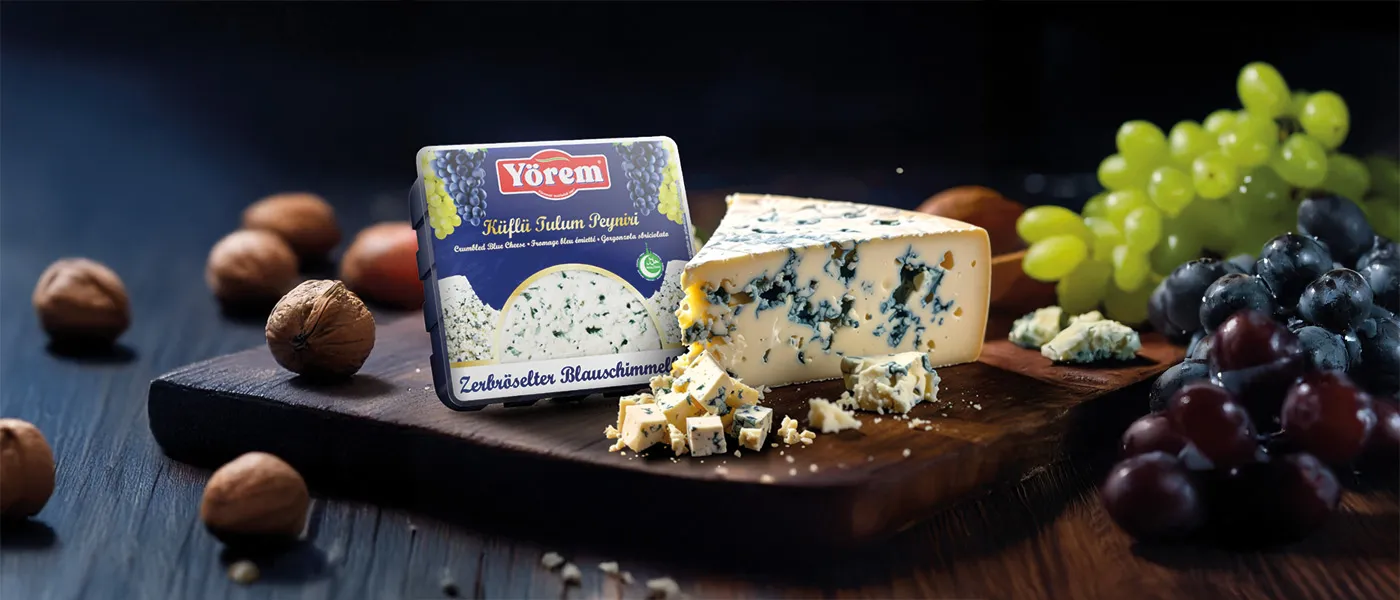 Eine Packung Yörem zerbröselter Blauschimmelkäse steht auf einem dunklen Holzbrett neben einem großen Stück Blauschimmelkäse mit sichtbaren blauen Adern. Vor dem Käse liegen kleine Würfel und Krümel. Um das Brett herum sind Walnüsse sowie grüne und dunkle Trauben dekorativ angerichtet.
